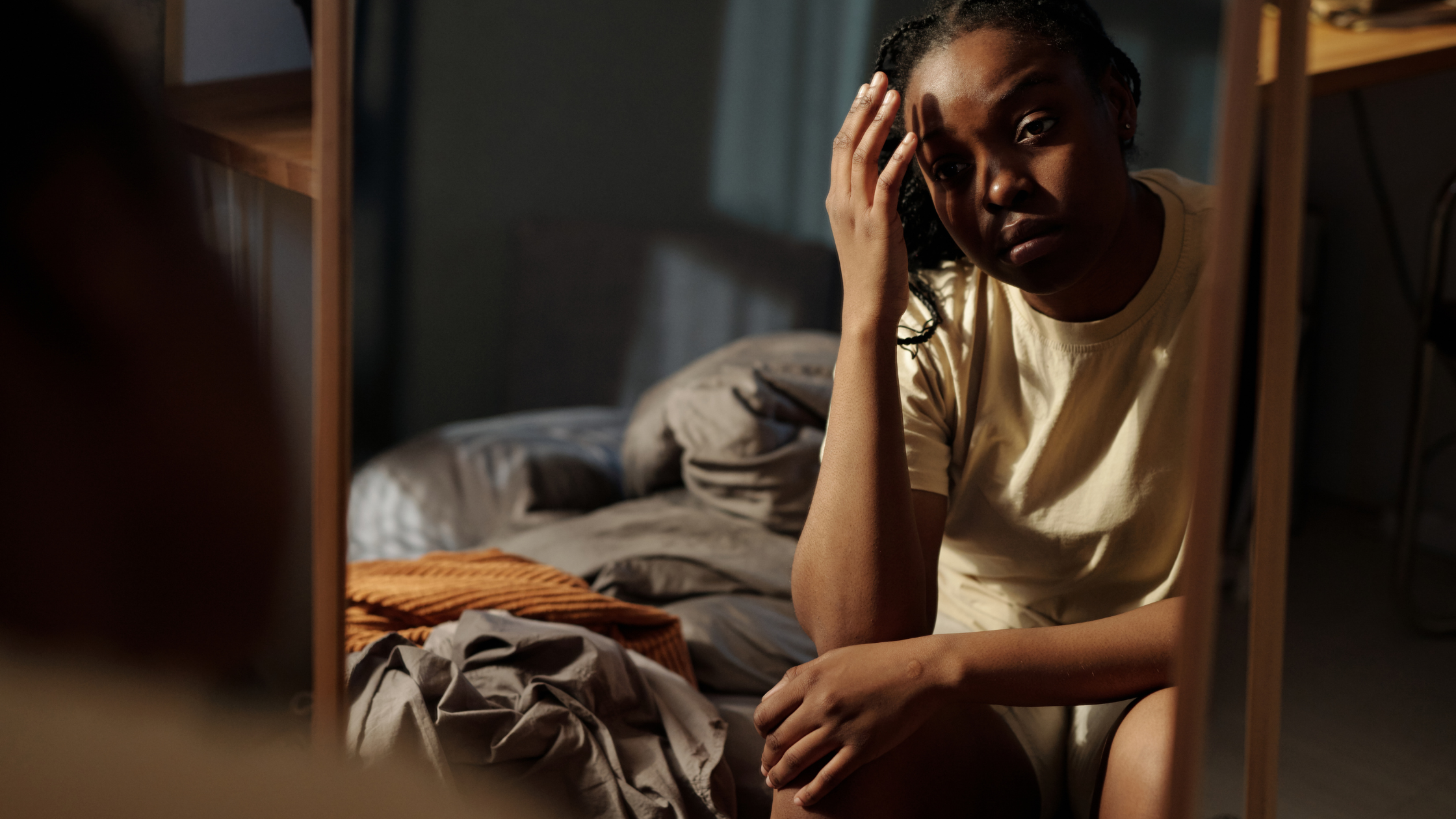 A woman sits on the edge of her bed looking tired and staring into a mirror